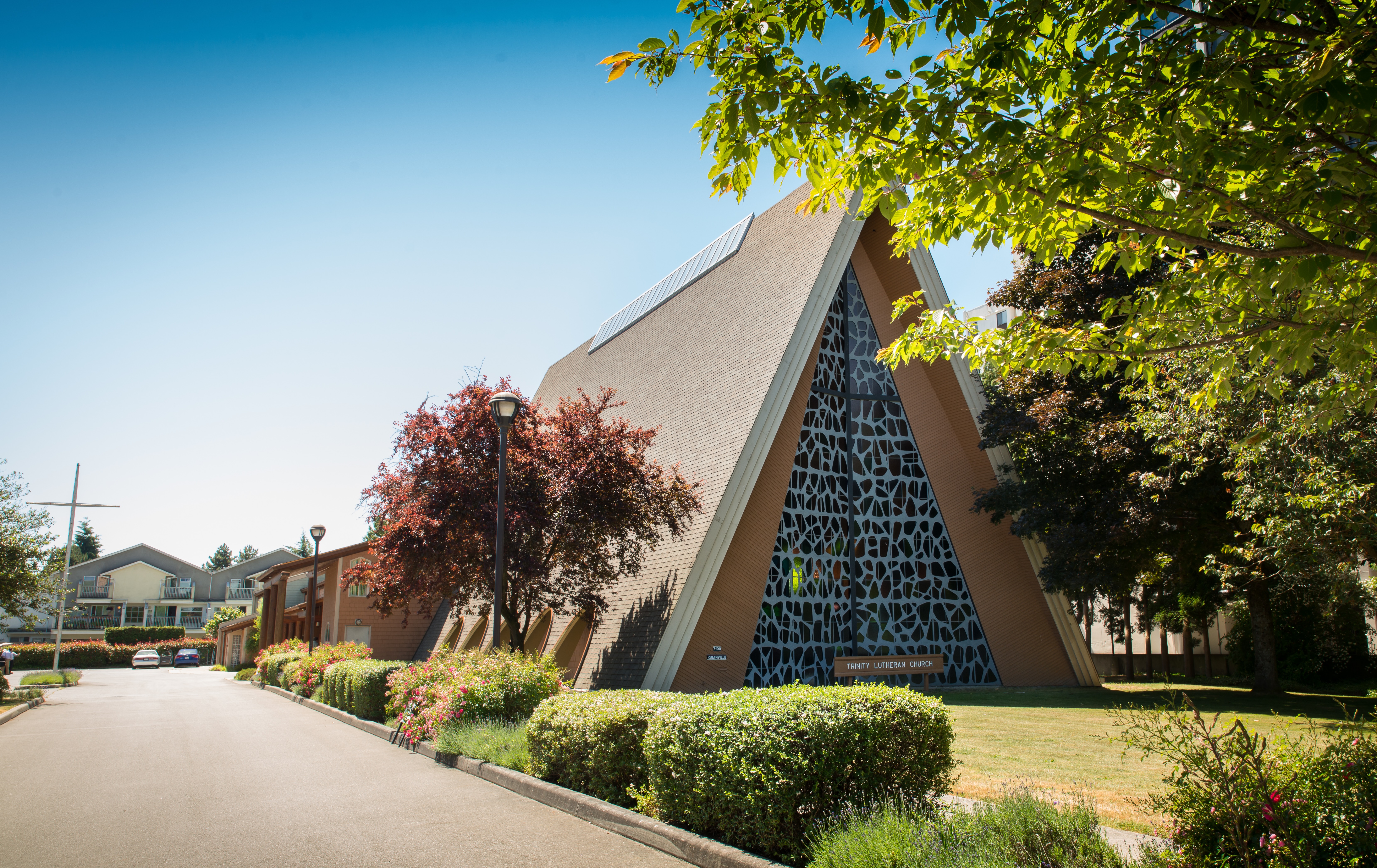 Exterior view of Trinity Lutheran Church in Richmond, BC, photographed by Tharaka Mapalagama. The image features the church's unique A-frame architecture, floor-to-ceiling stained glass mounted on a steel cross at the main entrance off Granville Avenue